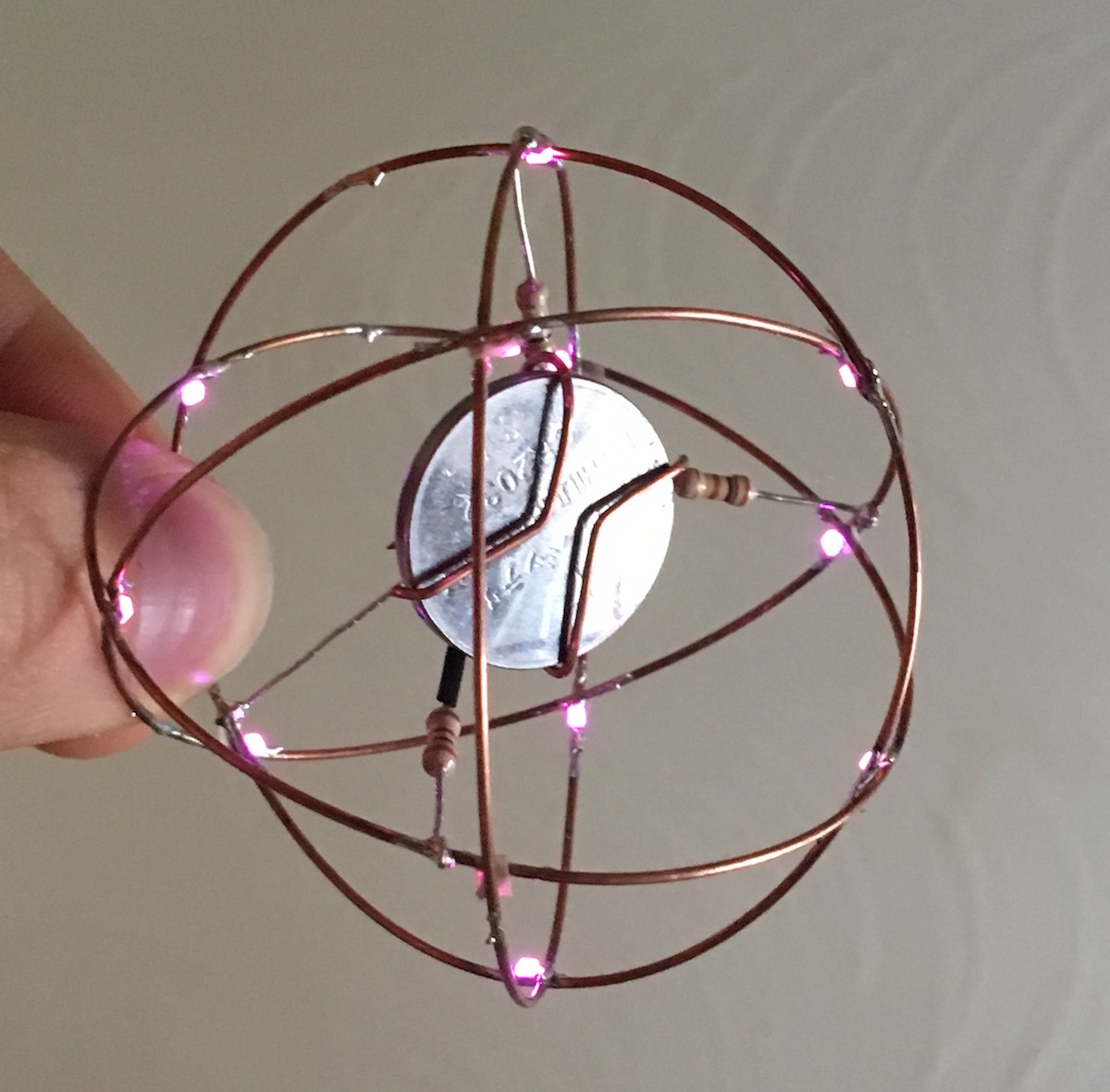 small metal sculpture made of four circular rings connected with glowing pink LEDs. a coin-cell battery is suspended in the centre by resistors. It's a glowy sparkly ball and here's another hint, this project goes in a touch-sensing interactive lil friend direction :)